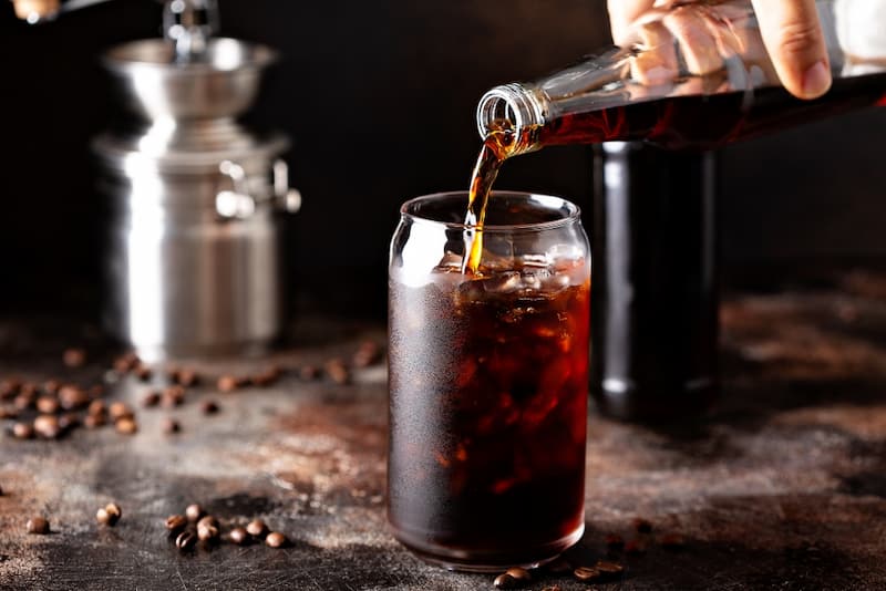 du café glacé est versé dans un verre à partir d'une bouteille, avec des grains de café et un moulin en arrière plan.