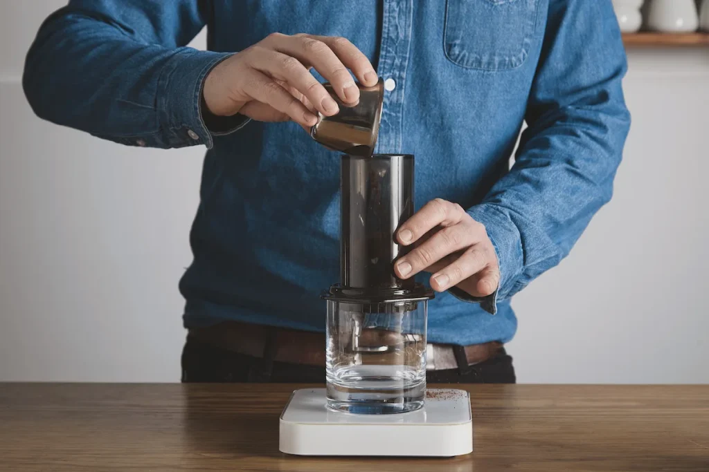 step by step aero press coffee preparation barista blue jeans shirt pours ground coffee powder from steel cup aeropress professional coffee brewing cafe shop (1)