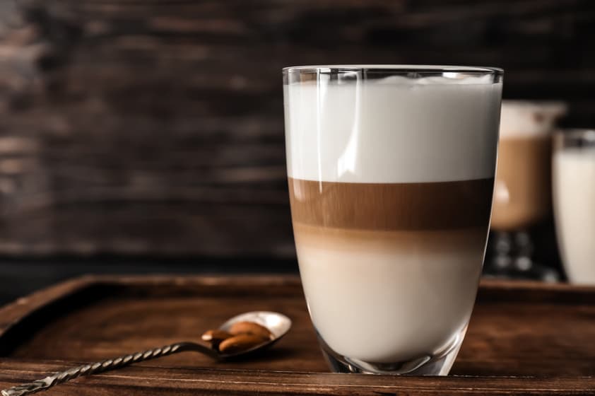un verre transparent rempli d'une boisson au café en couches, montrant des couches distinctes de lait, de café et de mousse, placé sur un plateau en bois avec une cuillère à côté.