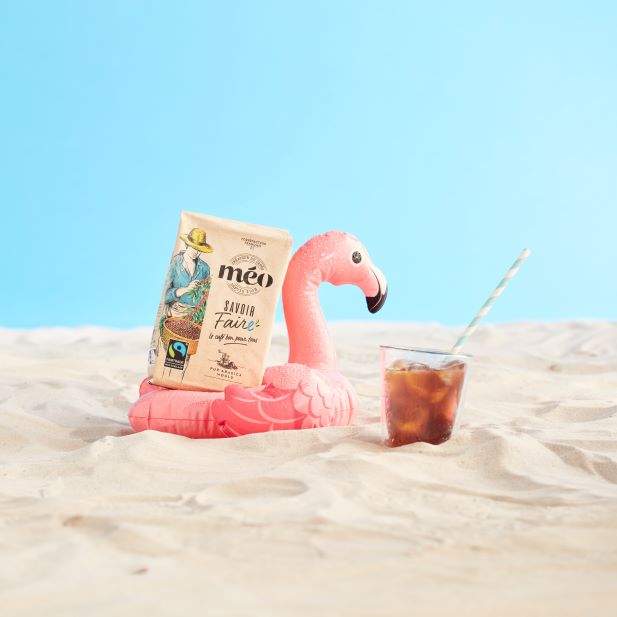 un flotteur de piscine flamant rose accueille un sachet de café méo. a côté se trouve un verre de café glacé avec une paille rayée, le tout situé sur une plage de sable sous un ciel bleu clair.