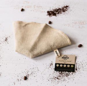 un filtre à café en tissu réutilisable repose sur une surface claire avec du marc de café et des grains dispersés. une petite étiquette avec le logo et les informations est visible à côté du filtre.