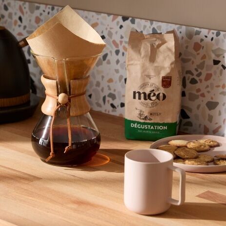 un comptoir avec une cafetière, un sac de café étiqueté « méo », une assiette avec des biscuits, une tasse rose, une bouilloire noire et des pots d'articles de cuisine en arrière plan.