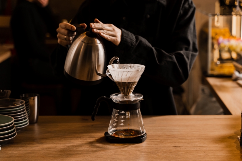 une personne verse de l'eau chaude d'une bouilloire dans une cafetière à filtre située au dessus d'une carafe en verre sur un comptoir en bois.