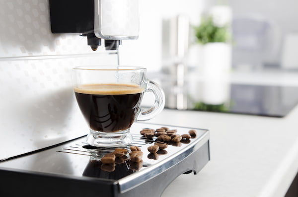 une tasse en verre transparent remplie de café noir est posée sur le bac d'égouttage d'une machine à café, avec des grains de café éparpillés tout autour.