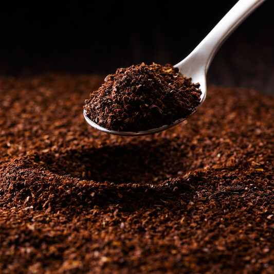 une cuillère en métal tient un petit monticule de café moulu au dessus d'un tas plus gros. le café est brun foncé et finement texturé sur un fond noir.