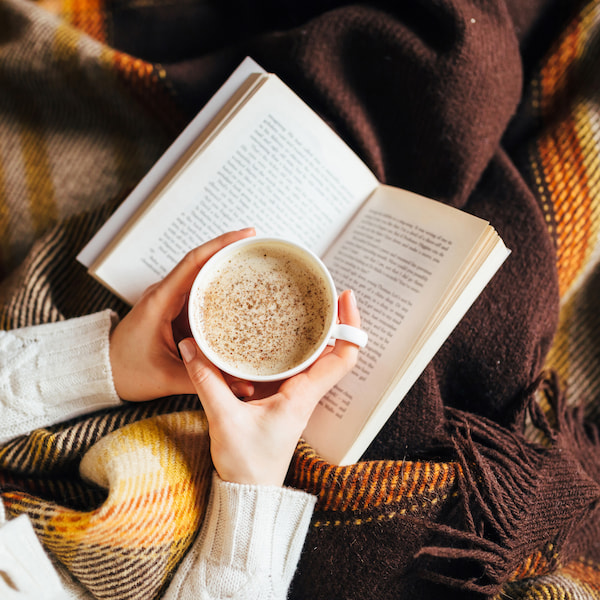 personne tenant une tasse de café, lisant un livre tout en étant enveloppée dans une couverture à carreaux.