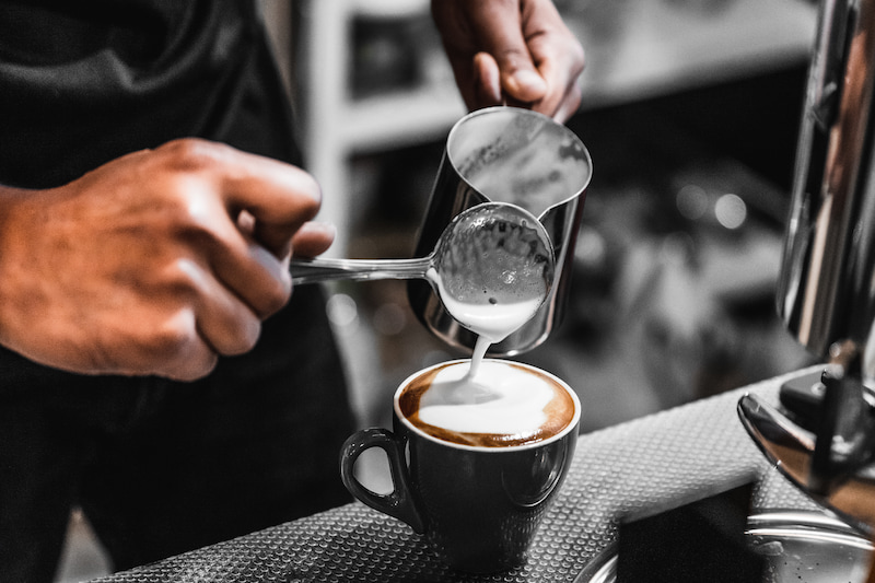 personne versant du lait cuit à la vapeur d'un pichet dans une tasse de café sur un comptoir.