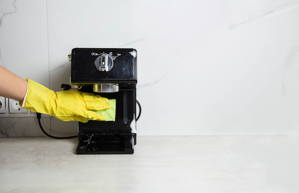 une personne portant des gants en caoutchouc jaune nettoie une machine à expresso noire avec une éponge sur un fond blanc.