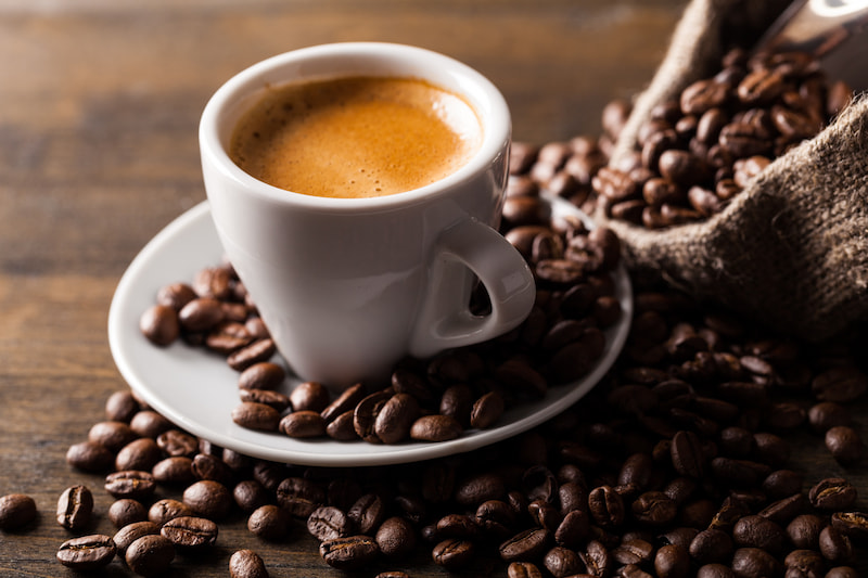 une tasse à expresso blanche sur une soucoupe entourée de grains de café éparpillés sur une surface en bois, avec un sac en toile de jute déversant plus de grains en arrière plan.