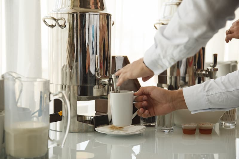 une personne portant une chemise blanche verse du café d'un distributeur dans une tasse blanche, avec des contenants de lait et de condiments à proximité.