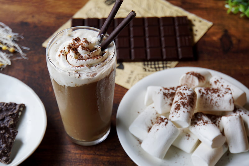 un verre de café glacé garni de crème fouettée et de bâtonnets de chocolat est posé sur une table en bois à côté d'une assiette de grosses guimauves saupoudrées de cacao. une barre de chocolat est partiellement visible en arrière plan.