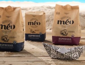 trois sacs de café expresso méo sont posés sur une surface en bois avec du sable et un fond de ciel bleu ; un petit bateau en papier est au premier plan.