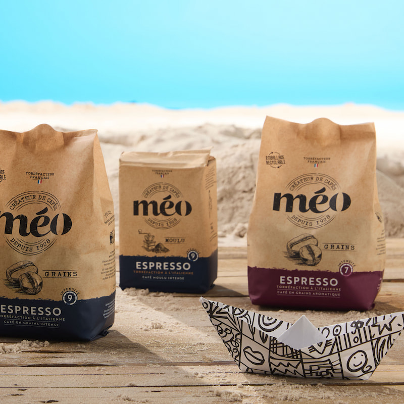 trois sacs de café expresso méo sont posés sur une surface en bois avec du sable et un fond de ciel bleu ; un petit bateau en papier est au premier plan.