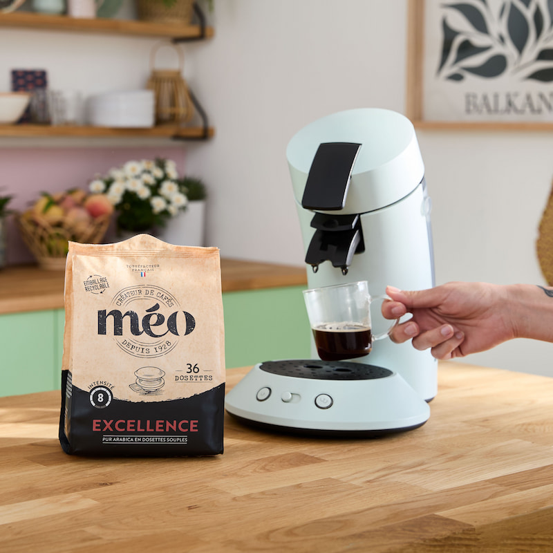 une personne tient une tasse en verre transparent sous une machine à café blanche, avec un sac de dosettes de café méo excellence sur le comptoir à proximité.
