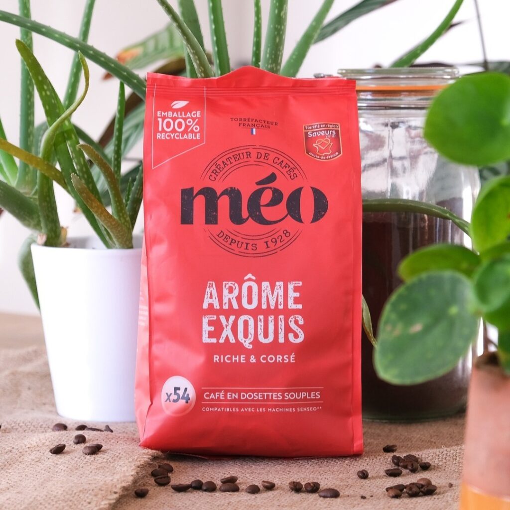 un sac rouge de café méo arôme exquis se trouve sur une table avec des grains de café éparpillés, près d'un pot de fleurs blanc, d'un bocal et de plantes vertes en arrière plan.