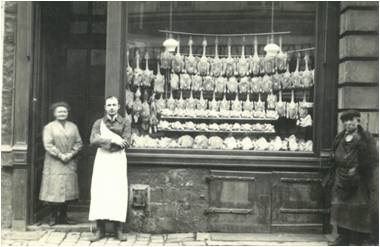 trois personnes se tiennent devant une boucherie avec de la volaille habillée accrochée à la vitrine et diverses viandes exposées sur le comptoir.