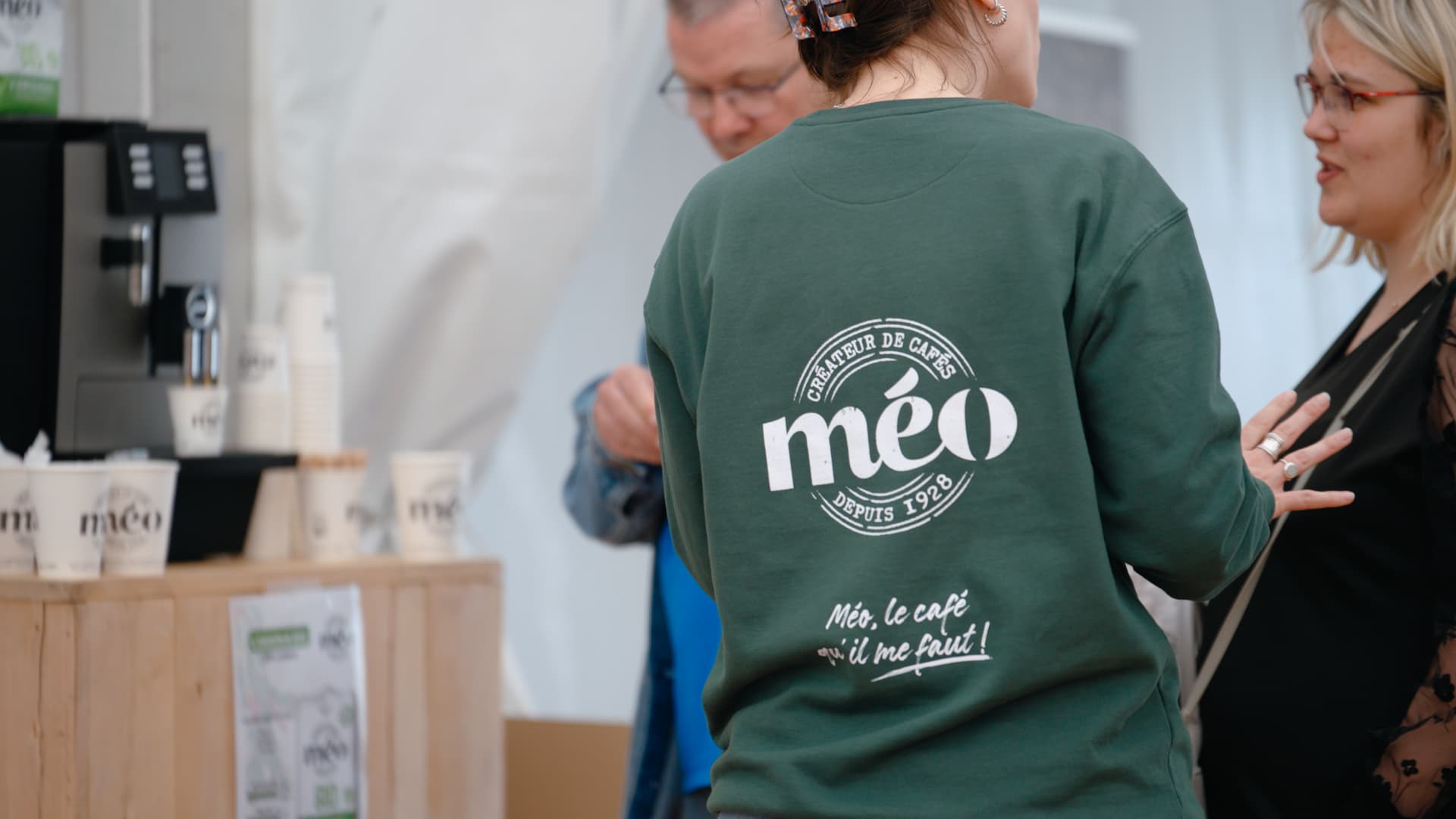 une personne portant un sweat shirt vert à café « méo » se tient à l'intérieur près d'un comptoir en bois avec des tasses à café et une machine à café, discutant avec deux autres personnes.