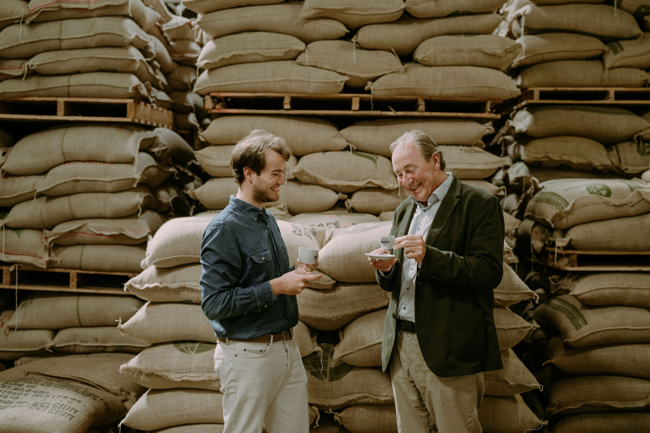 deux hommes se tiennent devant des sacs de jute empilés, souriant tout en tenant des tasses et des soucoupes, dégustant peut être du café ou du thé dans un entrepôt.