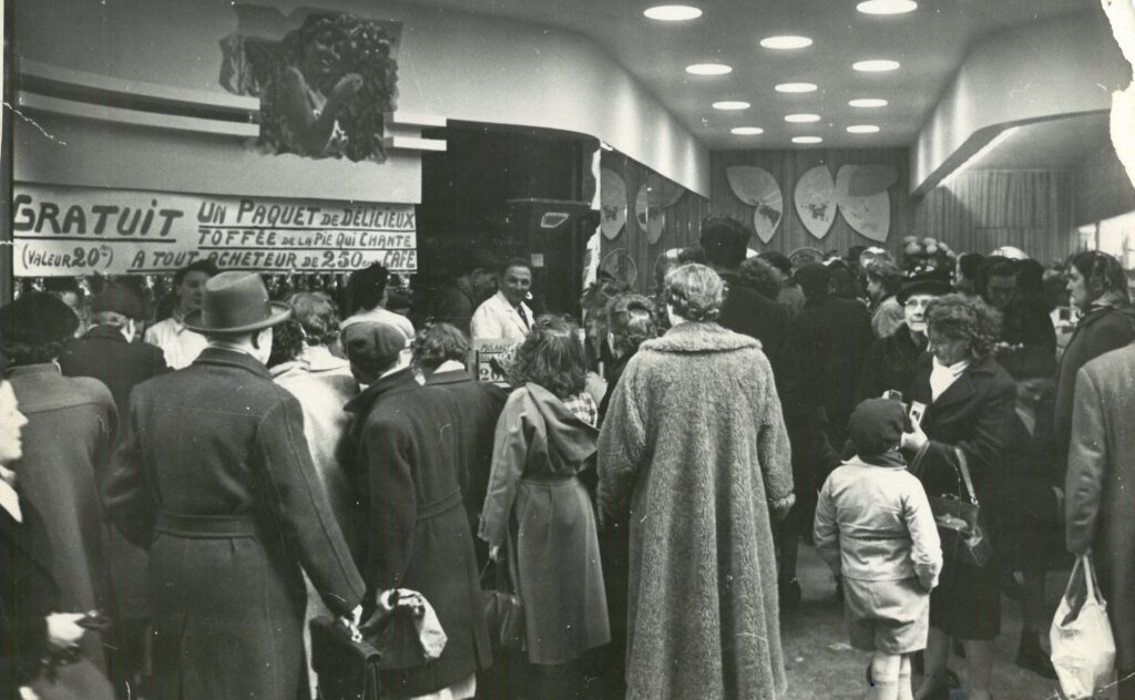 photo en noir et blanc de l'intérieur d'un magasin bondé, avec des gens en manteaux et chapeaux regardant un comptoir et une bannière faisant la publicité de caramels gratuits pour les clients.