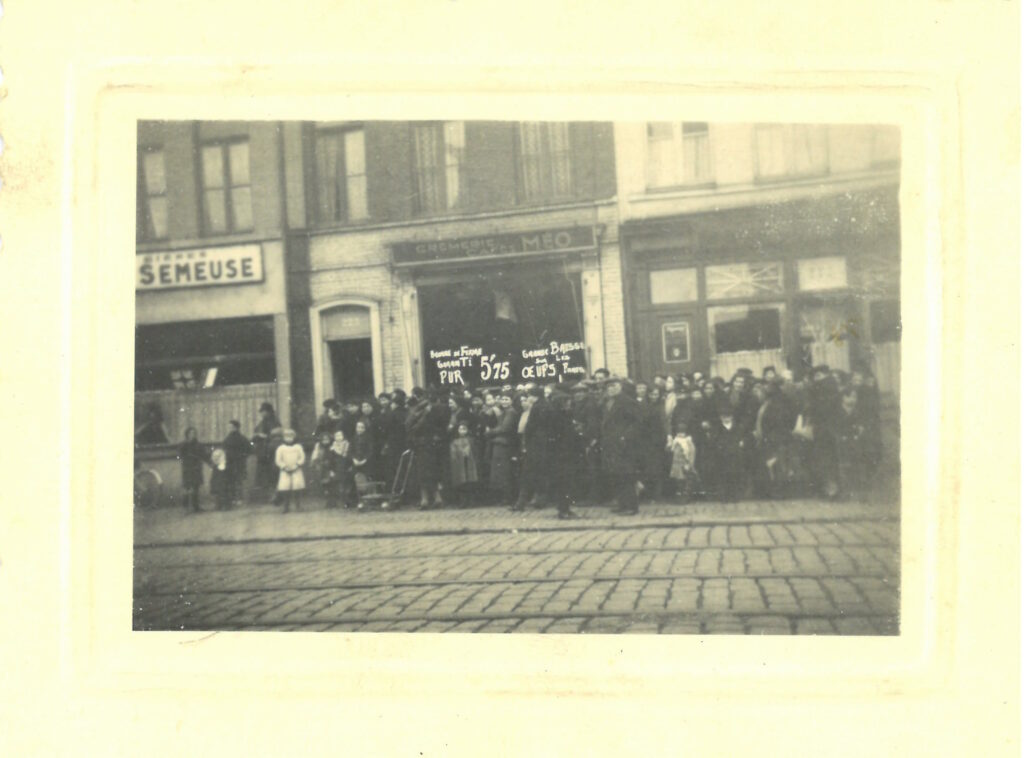 un grand groupe de personnes se tient dans une rue pavée devant des vitrines de magasins, tenant des pancartes indiquant les prix du sucre, du pain et d'autres produits.