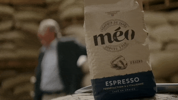 un sac de grains de café expresso méo est mis en évidence sur une surface tandis qu'un homme flou marche en arrière plan.