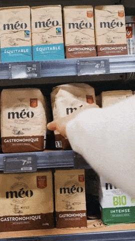 une personne prend un sac de café méo gastronomique sur une étagère de supermarché parmi d'autres emballages similaires.