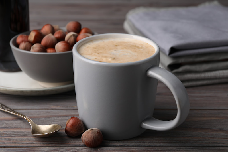 une tasse grise remplie de café mousseux est posée sur une table en bois à côté d'un bol de noisettes, d'une cuillère en argent et de serviettes grises pliées.