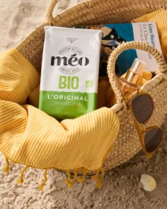 sac en paille sur le sable avec une serviette à rayures jaunes, des lunettes de soleil, un vaporisateur, un livre, un coquillage et un sachet de café bio méo bio à proximité.