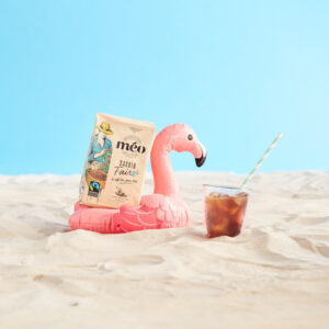 un sachet de café méo issu du commerce équitable est placé dans un porte boisson flamant rose gonflable sur le sable, à côté d'un verre de café glacé avec une paille rayée, le tout sur un fond bleu vif.