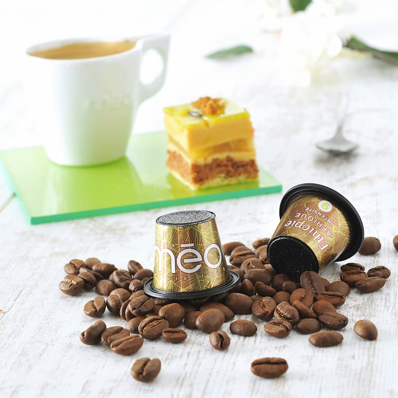 capsules de café et grains de café entiers sur une table, avec une tasse de café et un dessert en arrière plan.