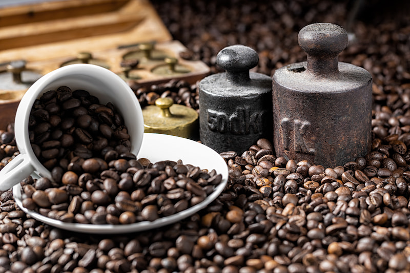 une tasse blanche renversée sur une soucoupe, renversant des grains de café torréfiés, avec plusieurs poids métalliques et plus de grains en arrière plan.