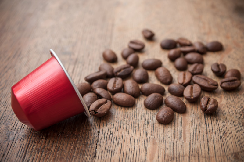 une capsule de café rouge est posée sur le côté, sur une surface en bois, et plusieurs grains de café sont éparpillés à proximité.