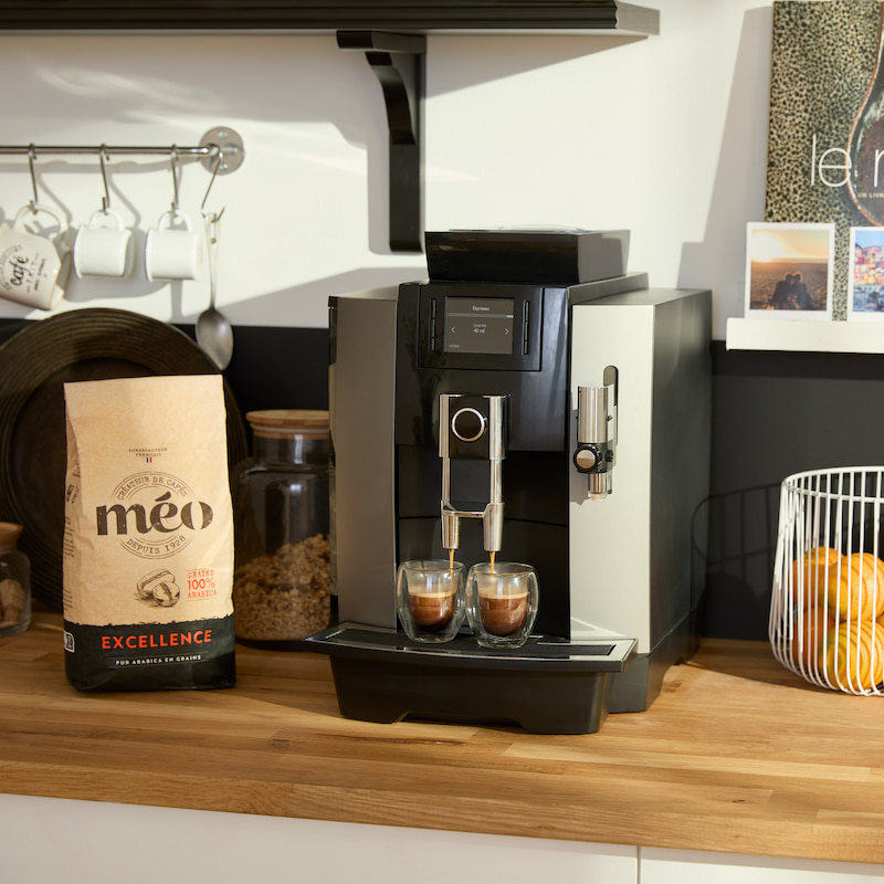 une machine à café prépare deux expressos sur le comptoir d'une cuisine, à côté d'un sac de grains de café méo, d'un bocal et d'un panier d'oranges.