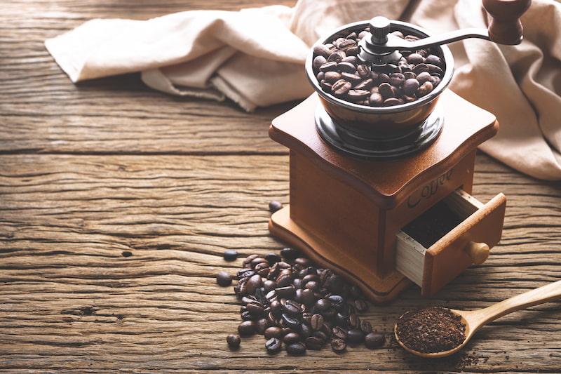 un moulin à café manuel avec des grains de café à l'intérieur, un petit tiroir avec du café moulu, des grains en vrac sur la table et une cuillère en bois avec du café moulu sur une surface en bois rustique.