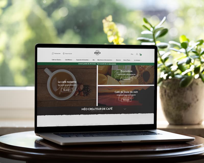 un ordinateur portable posé sur une table ronde affiche un site web sur le café ; une plante verte et une fenêtre avec une vue extérieure floue sont en arrière plan.
