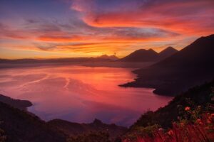 un coucher de soleil éclatant projette des teintes orange et roses sur un lac calme entouré de montagnes et de collines, avec des nuages épars dans le ciel.