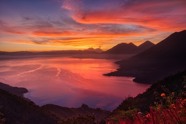 un coucher de soleil éclatant projette des teintes orange et roses sur un lac calme entouré de montagnes et de collines, avec des nuages épars dans le ciel.