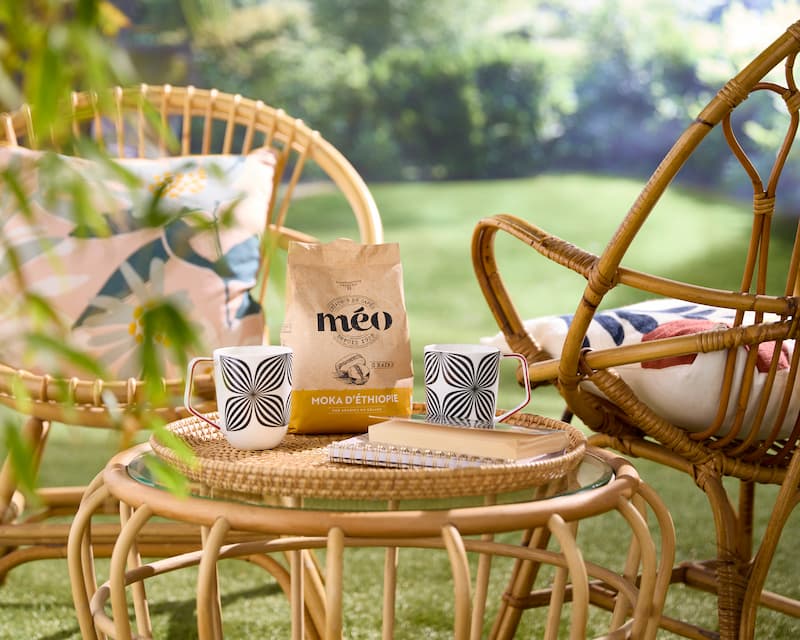 un ensemble de terrasse en rotin avec deux tasses, un sachet de café méo et un carnet de notes est posé sur une table en verre dans un jardin ensoleillé.