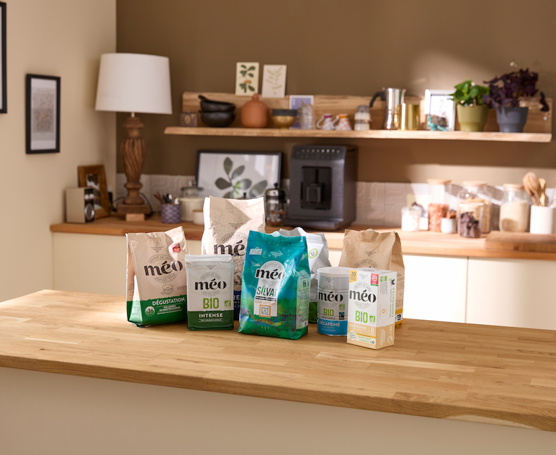 cinq paquets de produits de café méo sont présentés sur un comptoir de cuisine en bois avec des étagères, des plantes et des articles de cuisine en arrière plan.