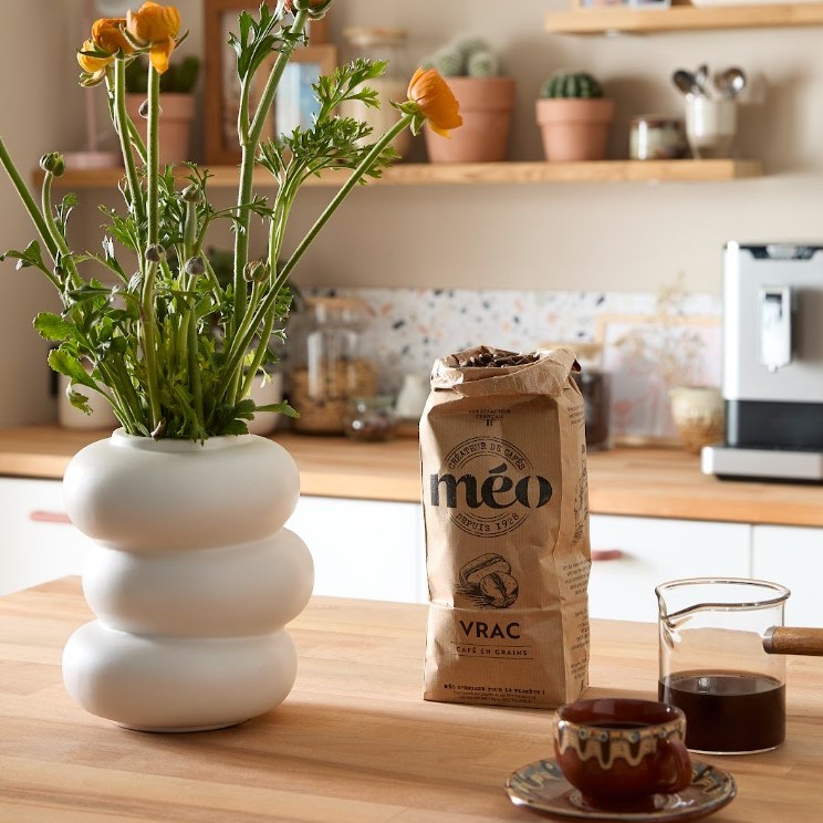 un sachet de café méo, un vase blanc avec des fleurs jaunes, une carafe de café en verre et une tasse sont posés sur un comptoir de cuisine en bois avec des étagères et une machine à café en arrière plan.