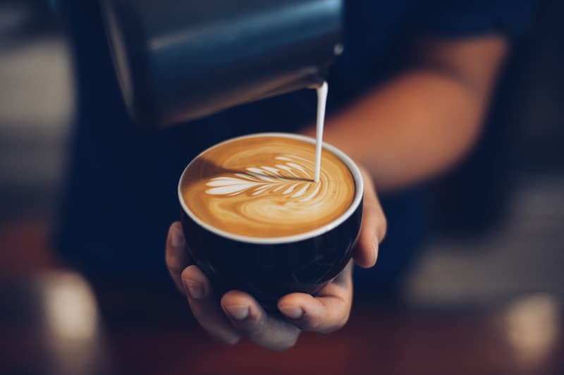 une personne verse du lait à la vapeur dans une tasse de café, créant ainsi un motif de latte art en forme de feuille sur la surface.
