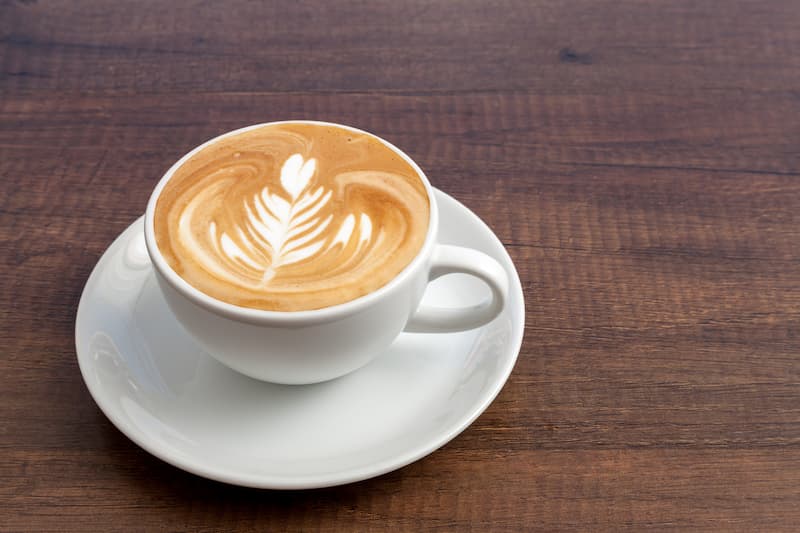une tasse de cappuccino avec un motif de feuilles dans la mousse est posée sur une soucoupe blanche sur une table en bois.