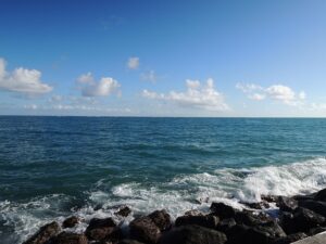 rivage rocheux avec des vagues qui s'écrasent et un océan bleu ouvert sous un ciel généralement clair avec des nuages blancs épars.