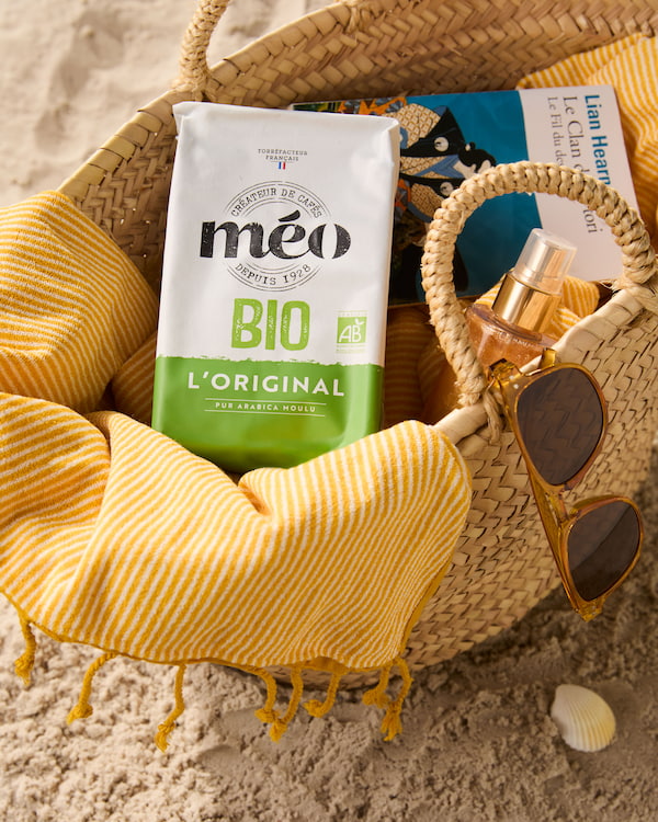 un sac de plage tissé sur le sable contient une serviette à rayures jaunes, un sachet de café méo bio, un livre, des lunettes de soleil et un petit vaporisateur.