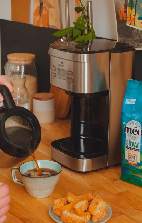 une personne verse du café d'une carafe en verre dans une tasse à côté d'une cafetière, d'un sachet de café et d'une assiette de biscotti sur un comptoir en bois.