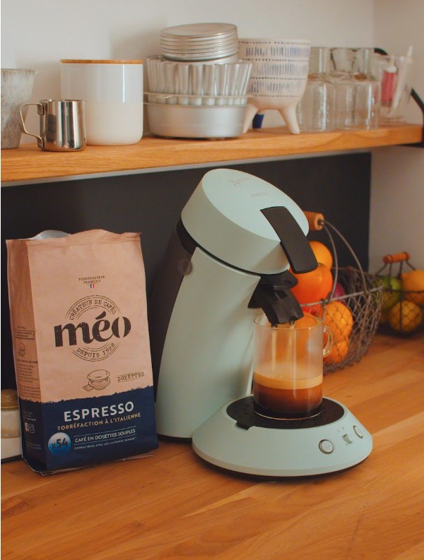 une machine à café bleu clair préparant un expresso dans un verre, avec un sachet de café expresso méo à côté, sur un comptoir de cuisine en bois.