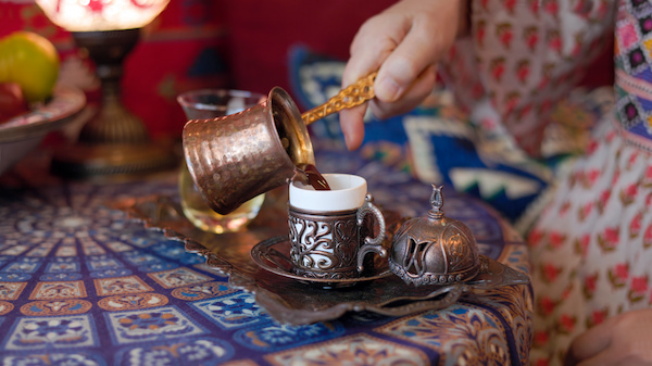 une main verse du café d'un pot en cuivre dans une tasse ornée sur un plateau décoratif, posé sur une nappe à motifs avec un plat couvert et un verre à l'arrière plan.