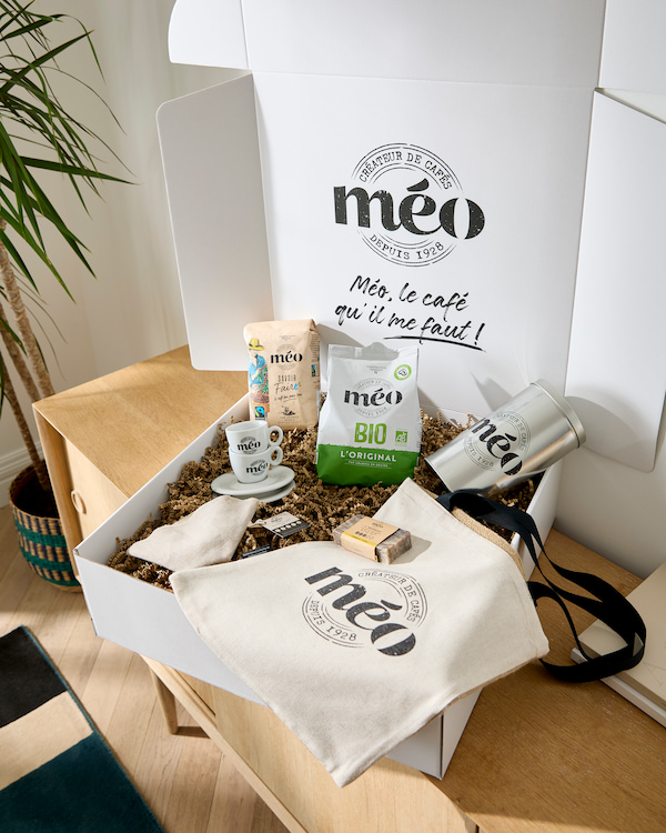 un coffret cadeau contenant des produits de café meo, un mug, une tasse et une soucoupe, un sac réutilisable, un gobelet et des petits sachets, disposés sur une table à côté d'une plante.