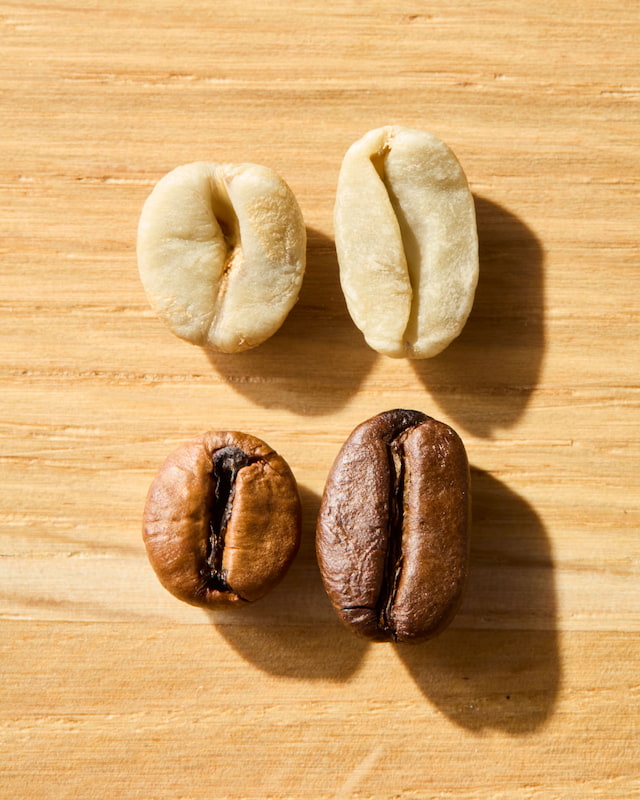 quatre grains de café sur une surface en bois ; deux sont crus et de couleur claire, et deux sont torréfiés et bruns, illustrant les différentes étapes de la transformation du café.