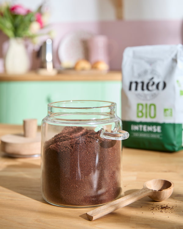 un pot en verre rempli de café moulu est posé sur un comptoir en bois à côté d'une pelle en bois, avec un sac de café biologique en arrière plan.